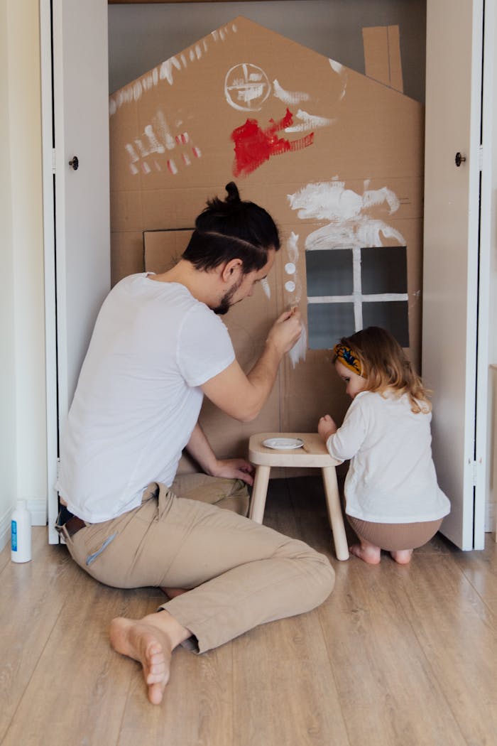 Mastering the First Impression: Your intriguing post title goes here Father and daughter painting a cardboard house indoors, enjoying quality time together.